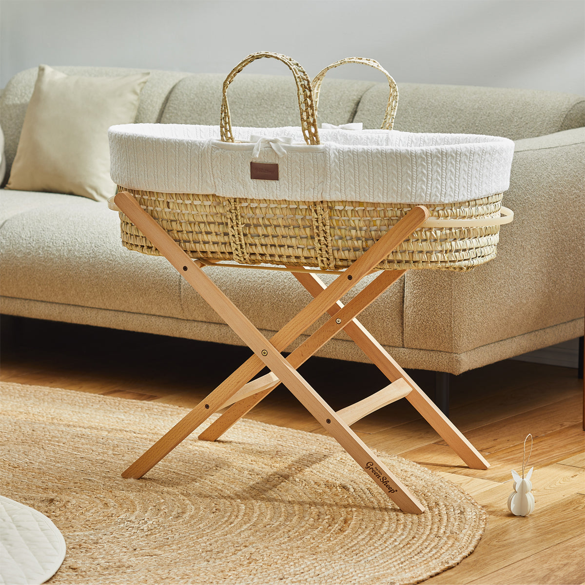 A baby's White Moses basket on a wooden stand, situated in front of a couch