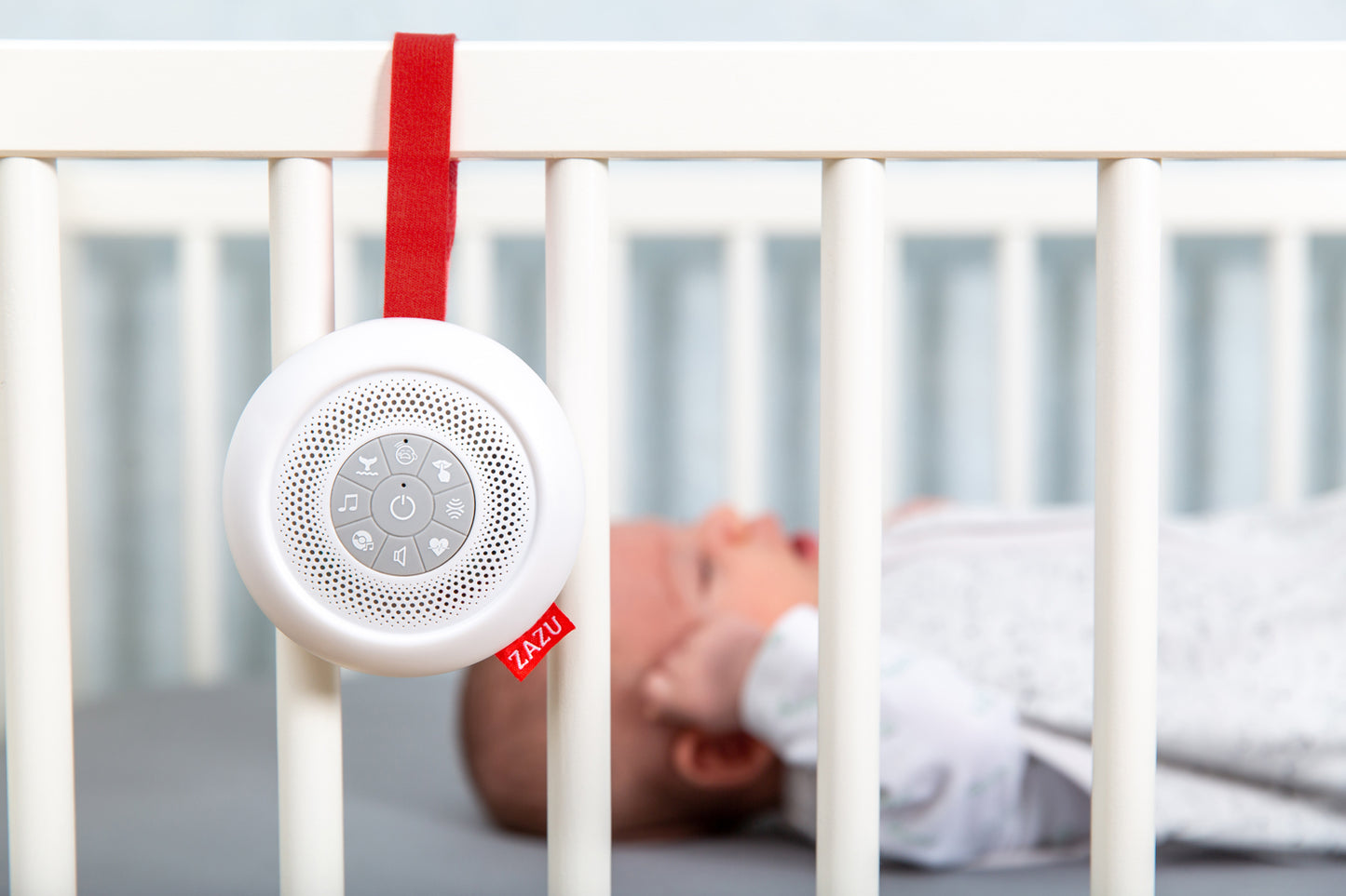 A round sound box with multiple feature buttons and red attachment strap, hanging on a cot bed while a baby sleeps.