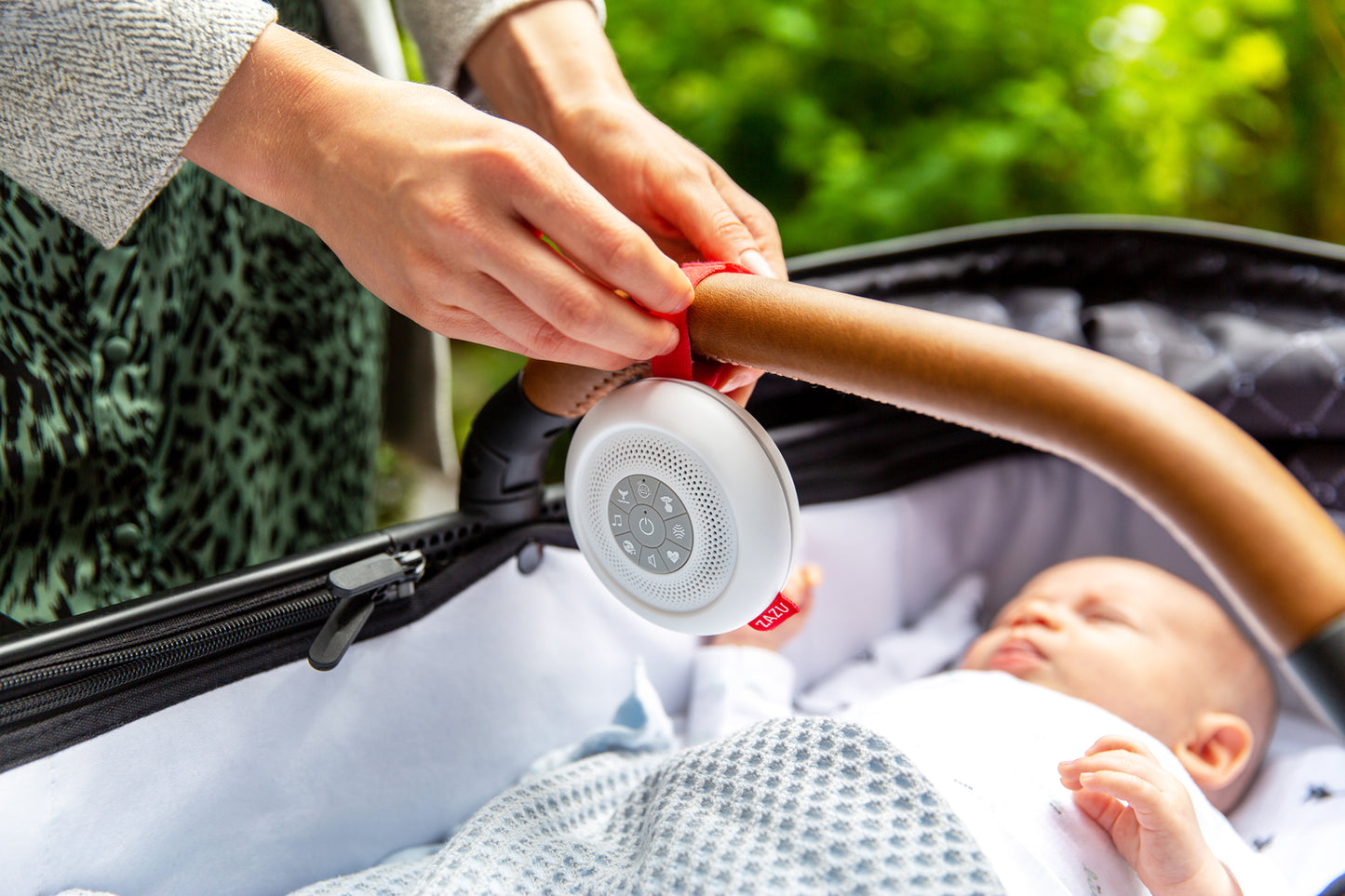 A round sound box with multiple feature buttons and red attachment strap, hanging on a carrycot bumper bar while a baby sleeps.