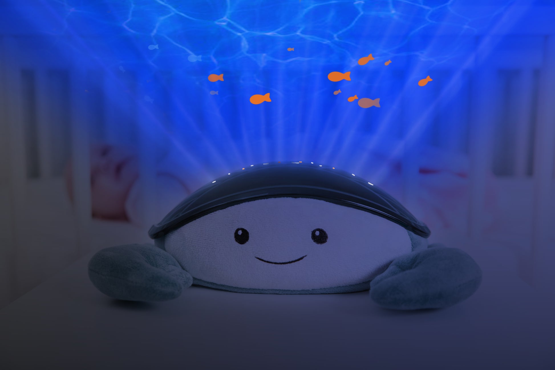 A teal and white crab shaped projector, sitting on a bedside locker displaying night light colours, while the baby rests in their cot.