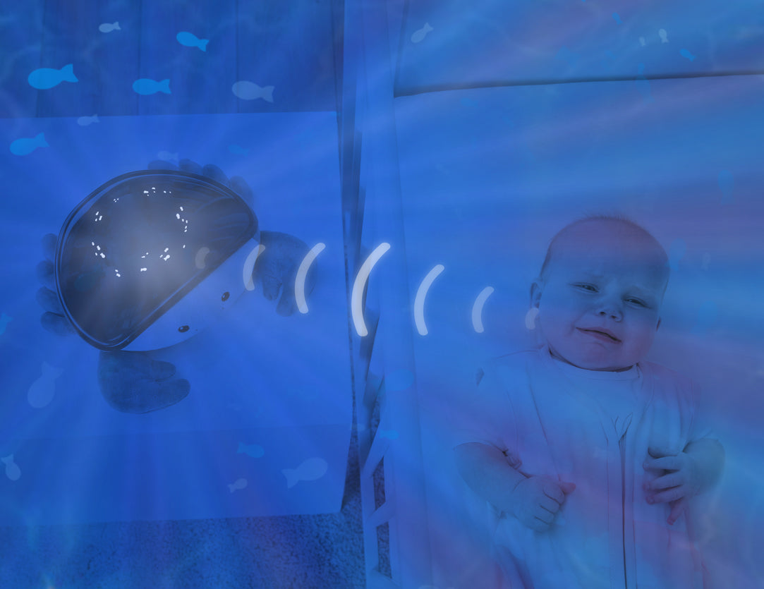 A teal and white crab shaped projector, sitting on a bedside locker displaying night light colours, while the baby rests in their cot.