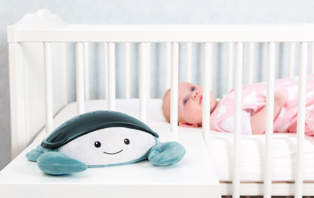 A teal and white crab shaped projector, sitting on a bedside locker while the baby rests in their cot.