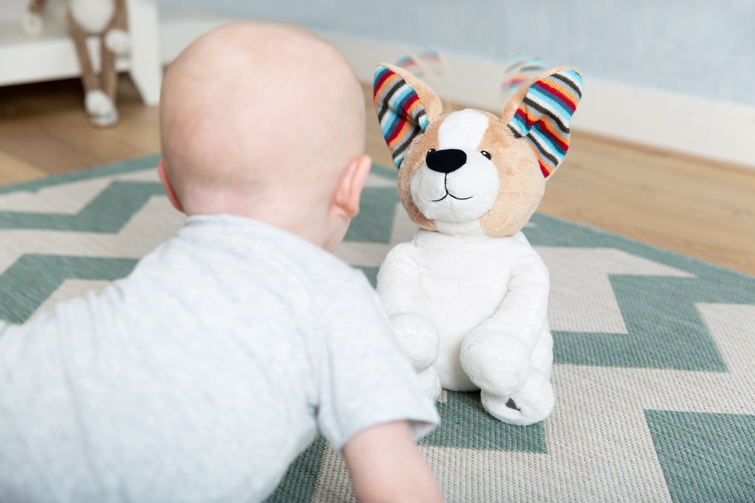 A tan and white shaped dog clapping toy, illustrating its clapping motion and sound, while a toddler plays with it on the floor.