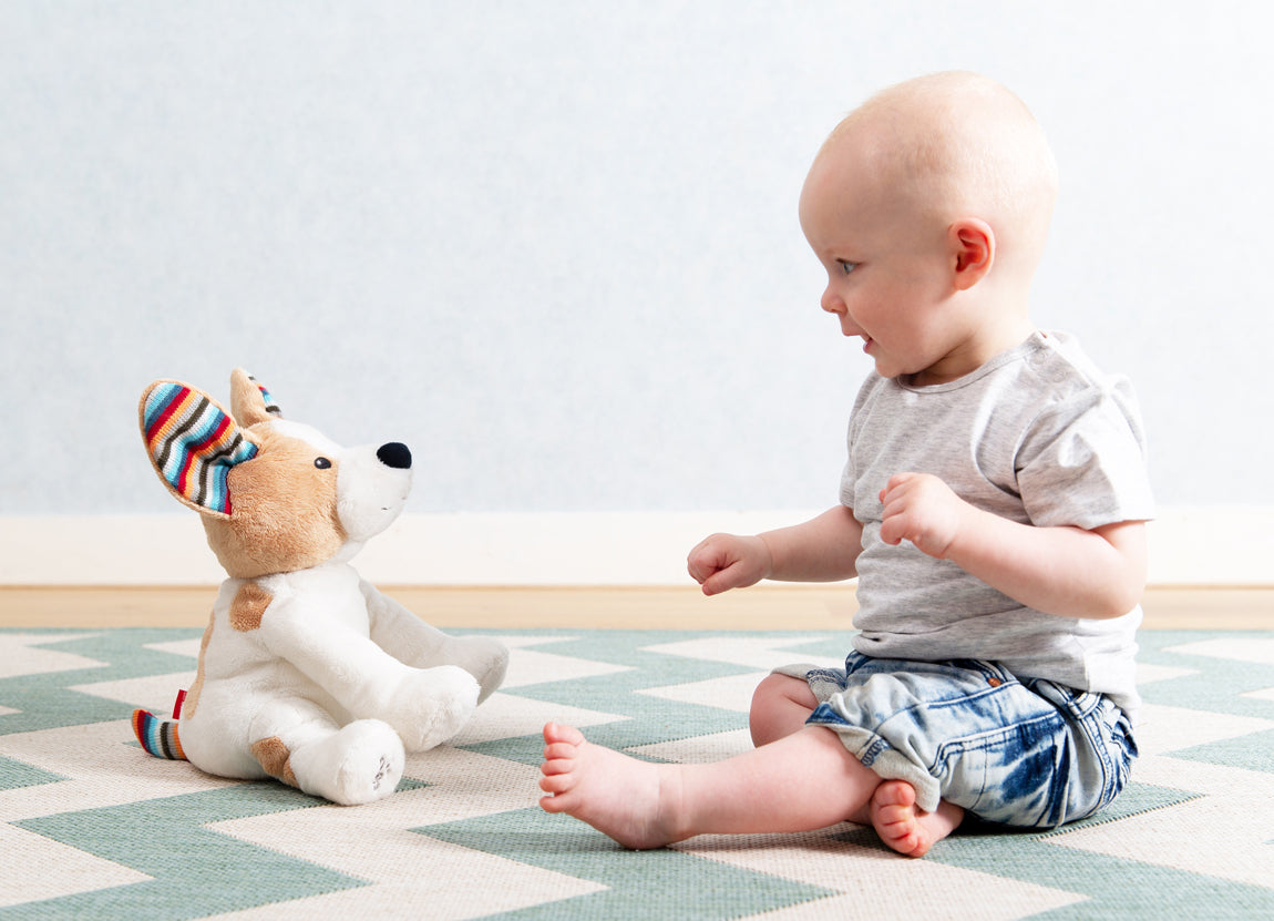 A tan and white shaped dog clapping toy, illustrating its clapping motion and sound, while a toddler plays with it on the floor.