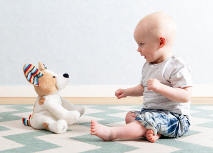 A tan and white shaped dog clapping toy, illustrating its clapping motion and sound, while a toddler plays with it on the floor.