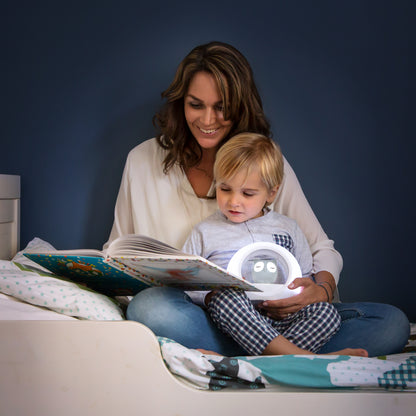 Mum and her toddler reading a book, while the toddle holds a grey and white round night light with an owl in the centre.