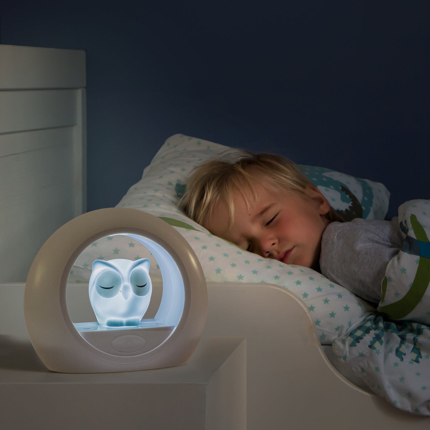 A toddler sleeps in their bed, and a grey and white round night light with an owl in the centre, sits on a bedside locker illuminated