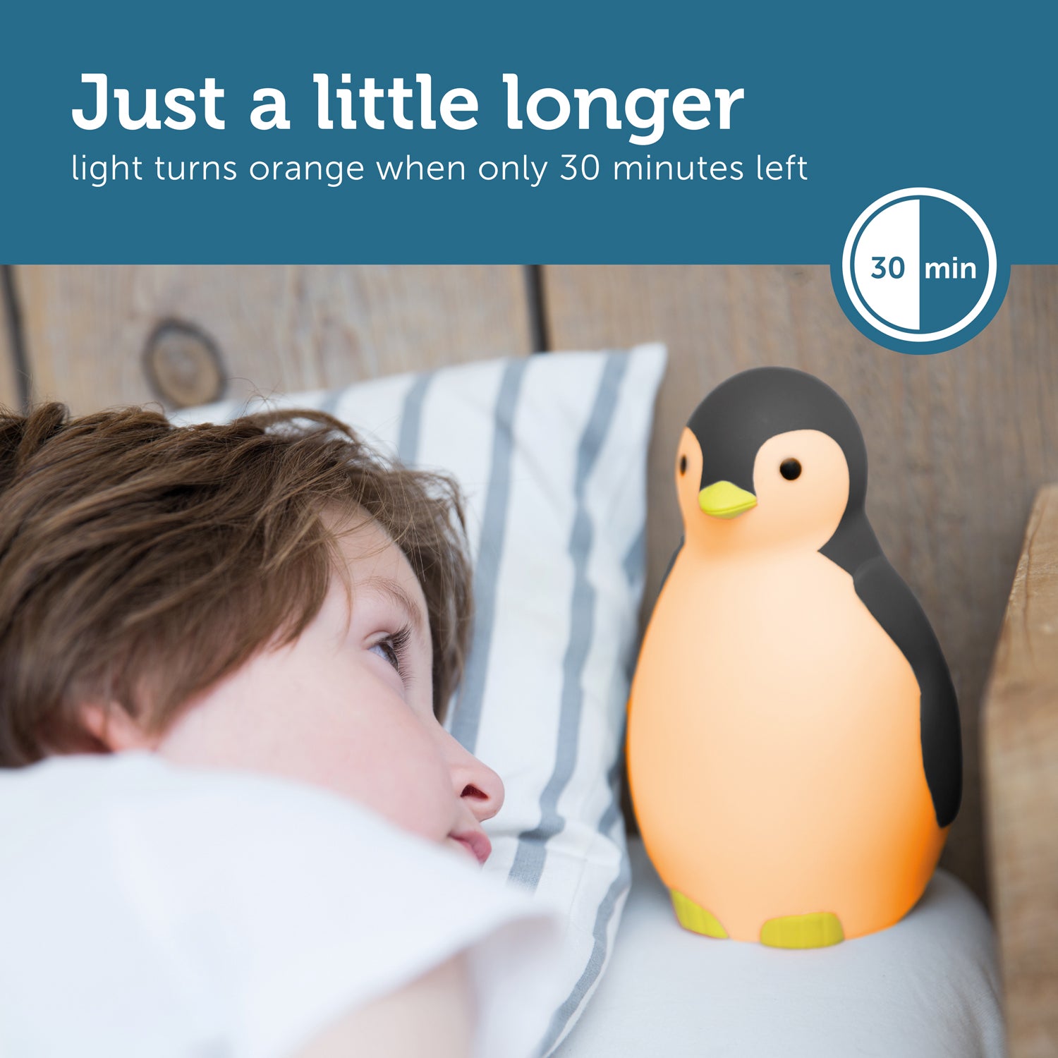 A grey and white musical penguin showing its changeable colours, while a toddler looks at it from their bed.