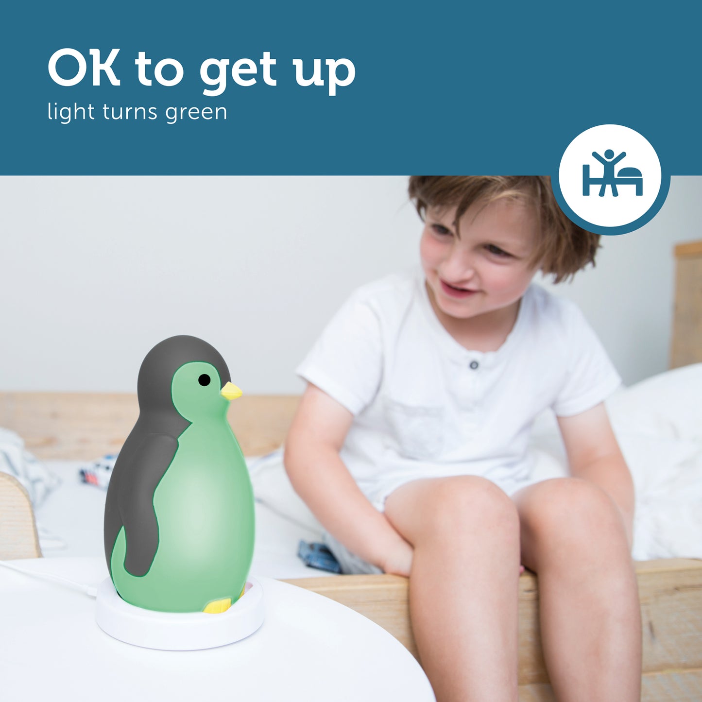 A grey and white musical penguin showing its changeable colours, while a toddler looks at it from their bed.