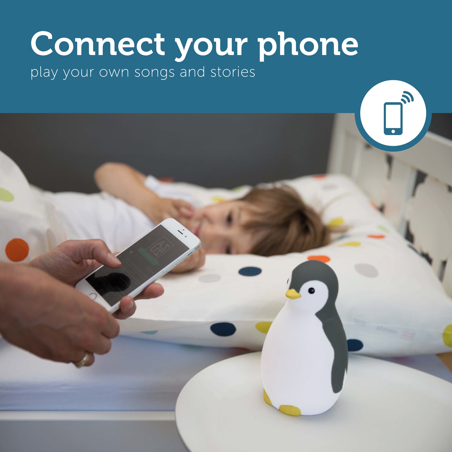 A grey and white musical penguin showing its changeable colours, while a toddler looks at it from their bed, as a parent uses the app on their phone to control the penguin's colours and sound.