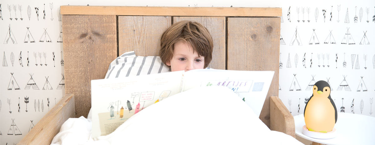 A grey and white musical penguin showing its changeable colours, while a toddler a toddler reads a book in their bed.