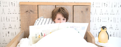 A grey and white musical penguin showing its changeable colours, while a toddler a toddler reads a book in their bed.