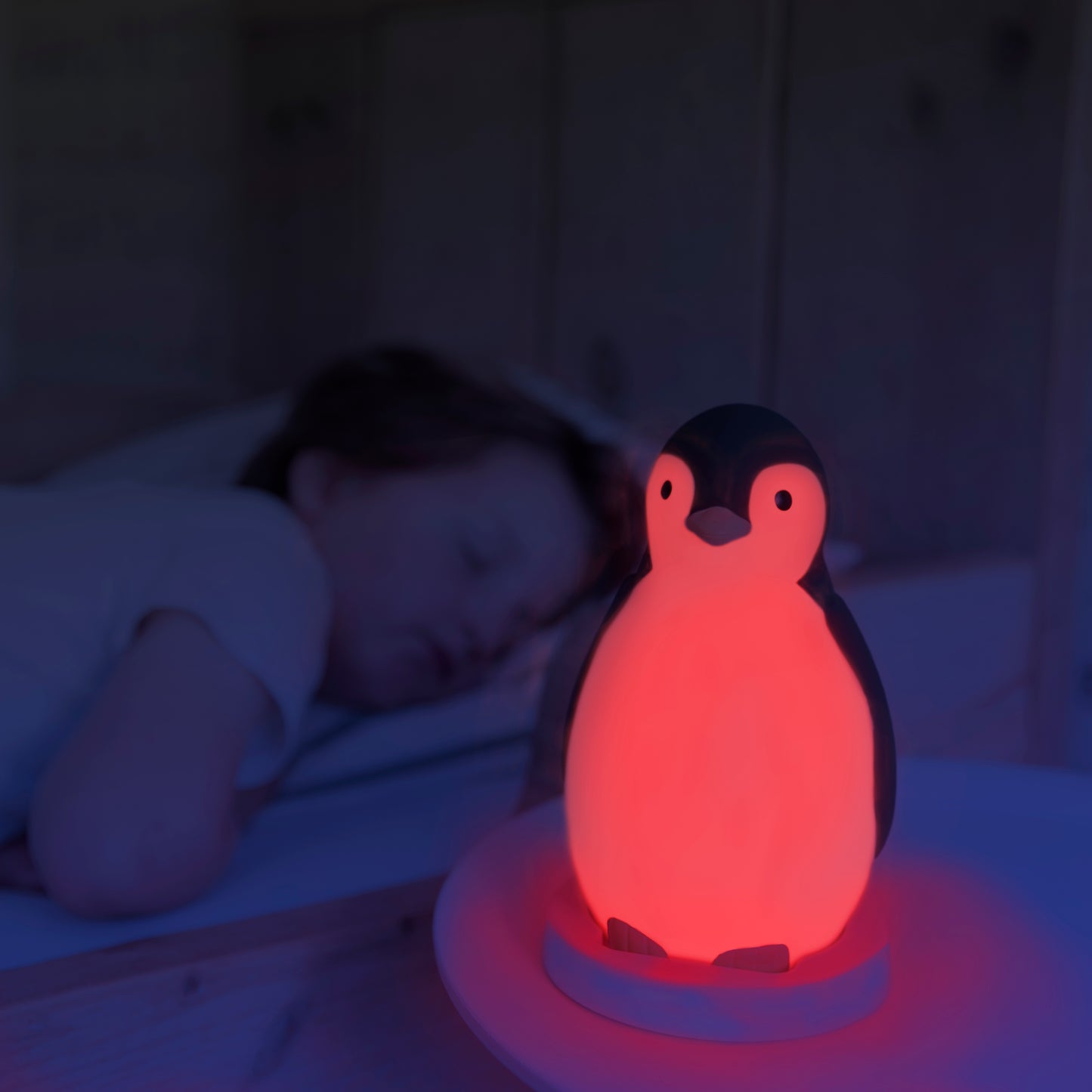 A grey and white musical penguin showing its changeable colours, while a toddler sleeps in their bed.