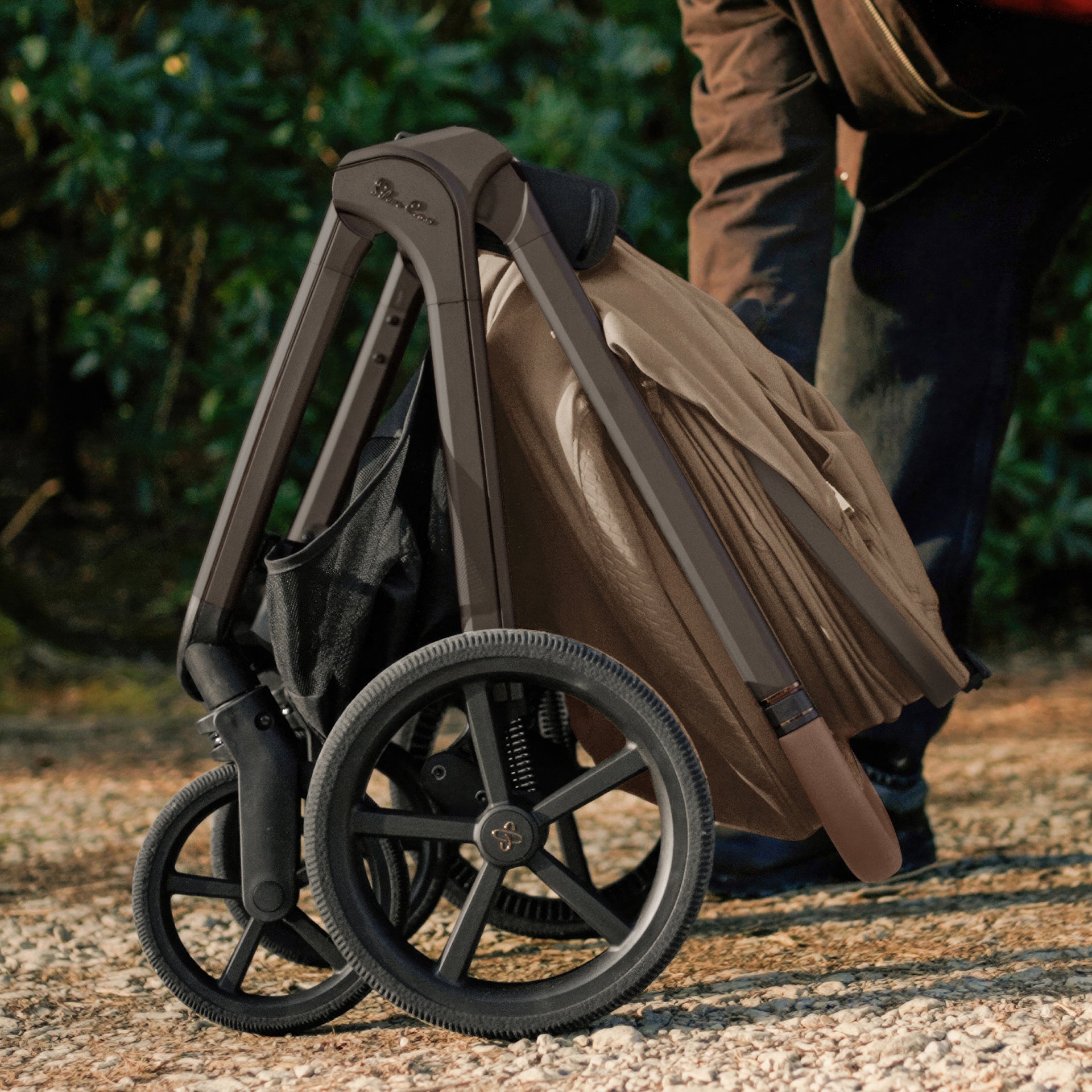 A multi-piece silver cross cove 2 travel system set in taupe brown and black, displayed on white pedestals against a white background. The set includes a stroller, bassinet, car seat in taupe, infant seat, footmuff, backpack, car seat liner, and accessories.