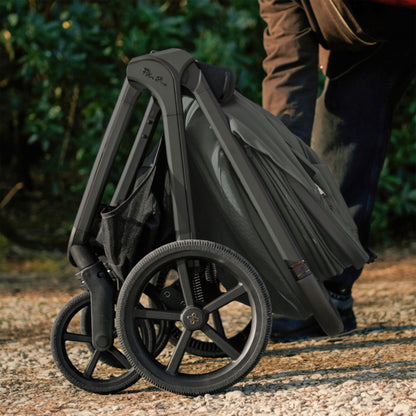 A multi-item silver cross cove 2 travel system set in dark olive green and black, displayed on white pedestals against a white background. The set includes a stroller, bassinet, car seat in black, infant seat, travel system cover, bassinet bag, car seat liner, and a brown backpack.