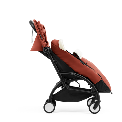 A stroller with a rust-colored padded footmuff, cream fleece lining, and a black frame, shown from the front on a white background.
