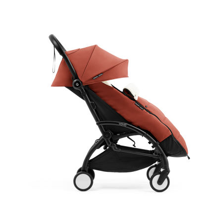 A stroller with a rust-colored padded footmuff, cream fleece lining, and a black frame, shown from the front on a white background.
