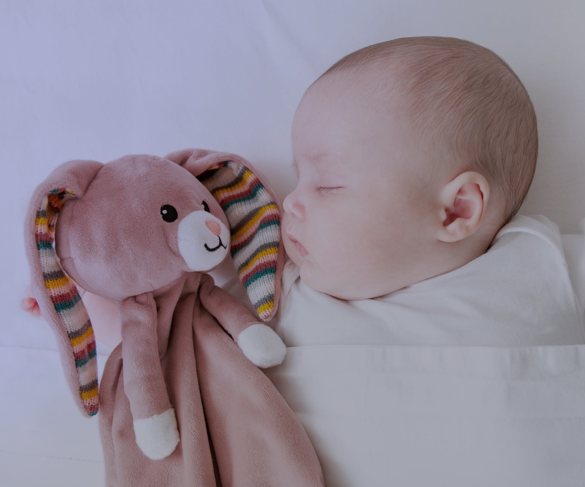 A pink bunny baby comforter resting beside a baby while the baby sleeps.