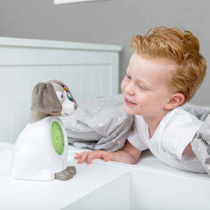 A beige and white dog with a clock in its colour changing belly, while a toddler looks at the sleep trainer on their bed.