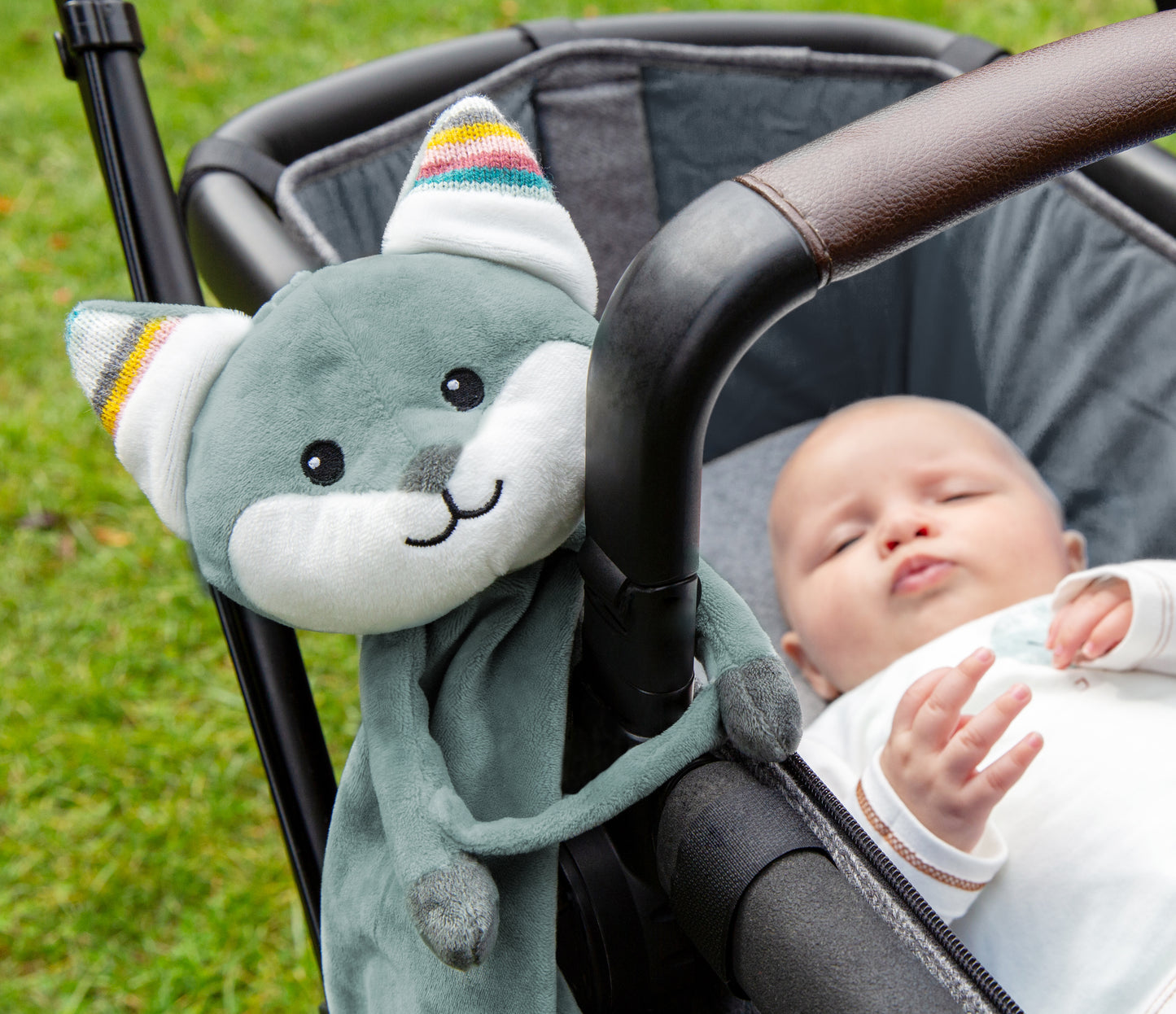 A teal fox baby comforter, attached to a pram carrycot bumper bar, while the baby sleeps in the carrycot.