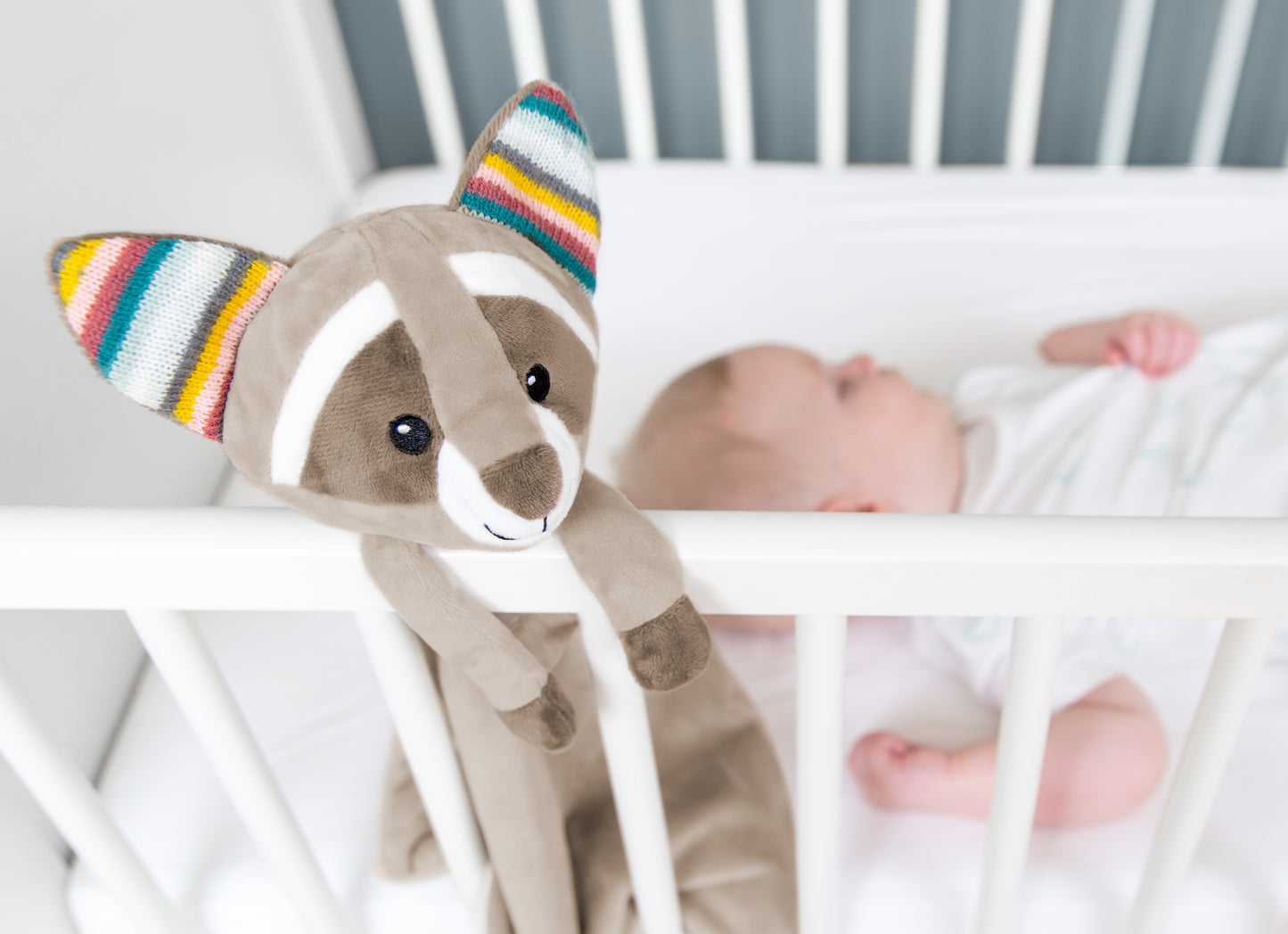 A beige racoon baby comforter attached to a cot bed while a baby sleeps.