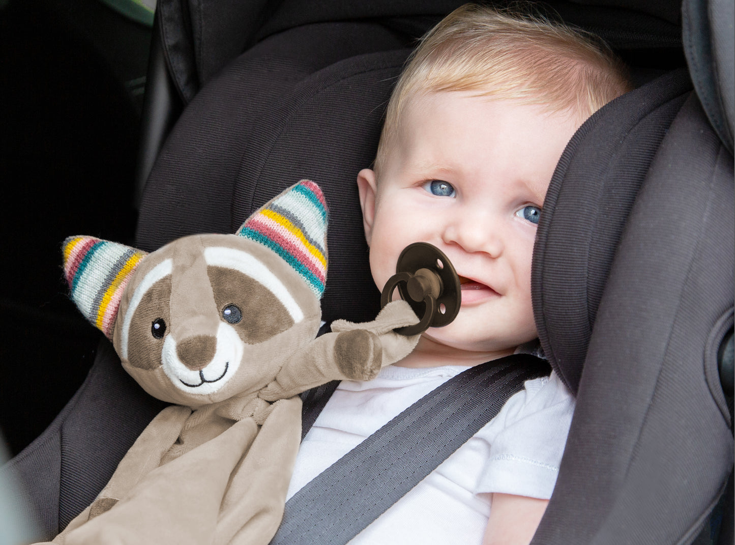 A beige racoon baby comforter lying a, while the baby sleeps, attached to a dummy, with a baby n a car seat.